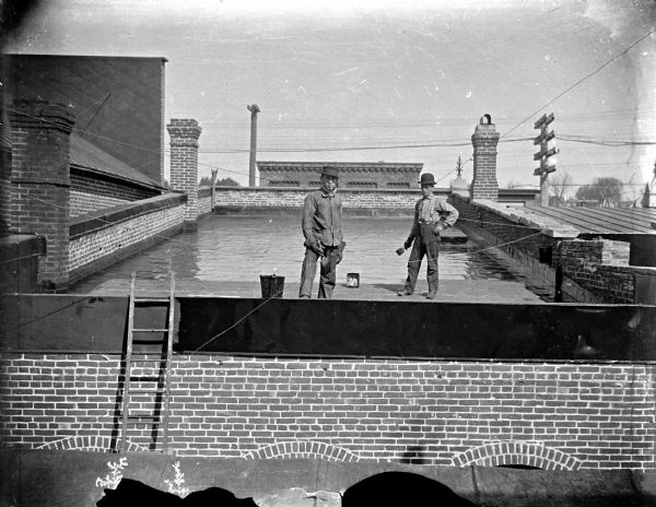 Outdoor view of two men standing on top of a building. Identified as probably the top of the Jackson County Bank from the photography studio of Van Schaick.