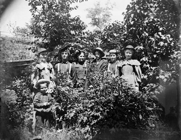 Outdoor group portrait of seven individuals, two young men, four young women, and a child, posing standing among vegetation.