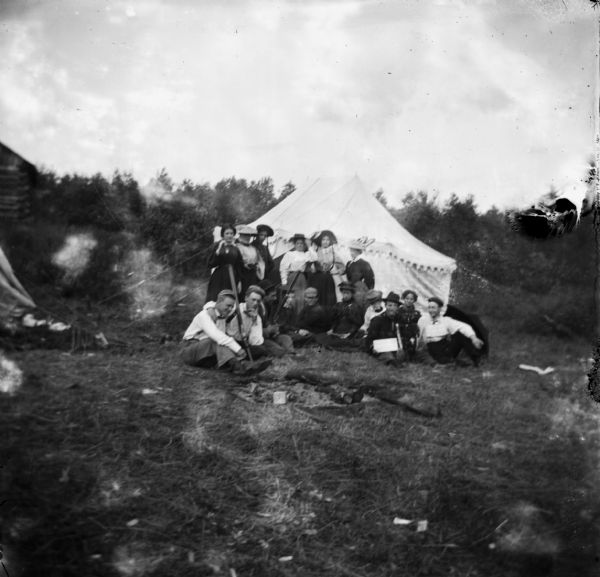 Outdoor view of fourteen men and women posing standing and sitting in front of a tent in a field.