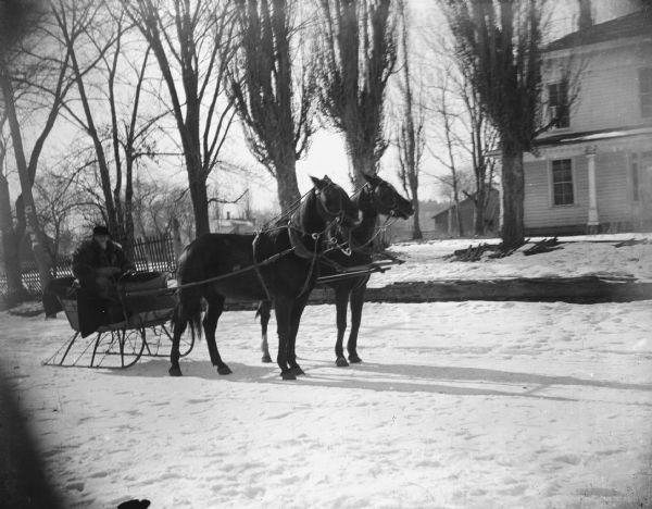 View towards a man posing sitting in a sleigh pulled by a team of two horses on a snow-covered street. There is a house in the background.