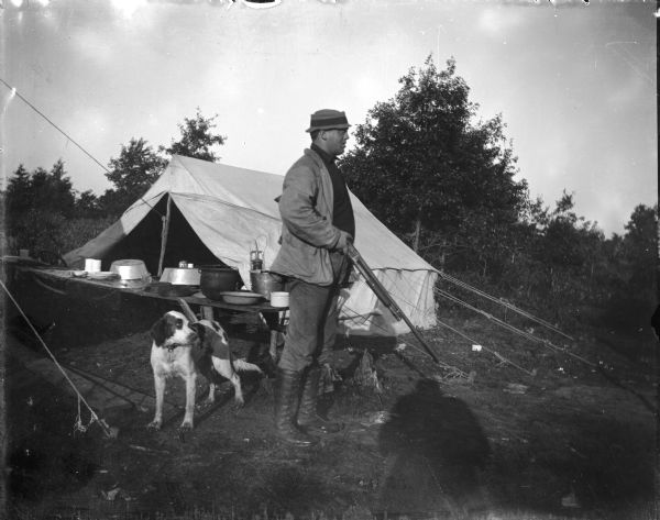 View towards a European American man posing standing and holding a gun. Behind him is a table and a tent, and behind him on the left is a dog.