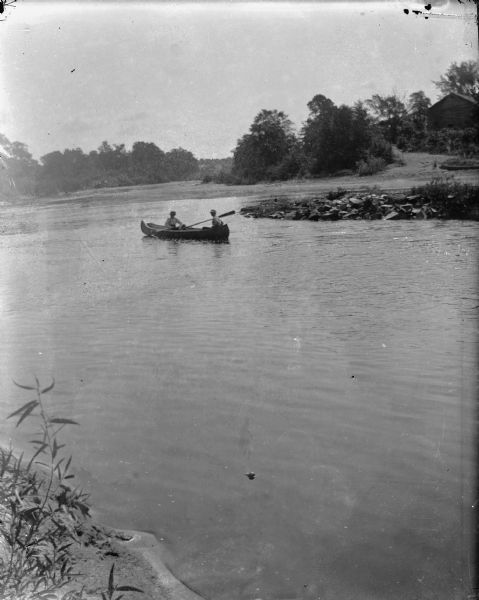 View from shoreline towards two people in a canoe on a river. There is a building on the far shoreline.