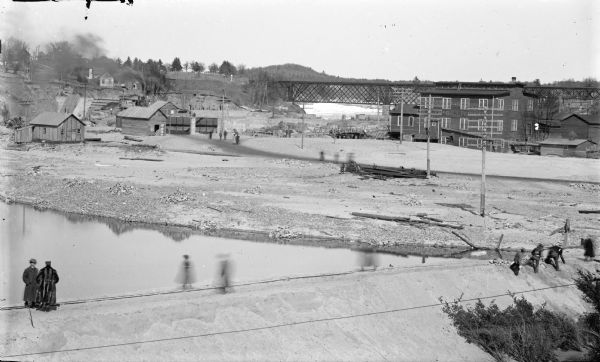 Elevated view of buildings, a railroad bridge, and in the foreground people standing on what may be a dam over the river.