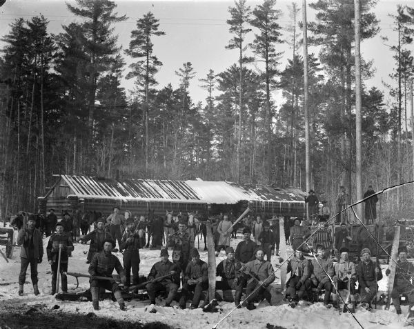 Outdoor group portrait of loggers with their tools near their lodge.