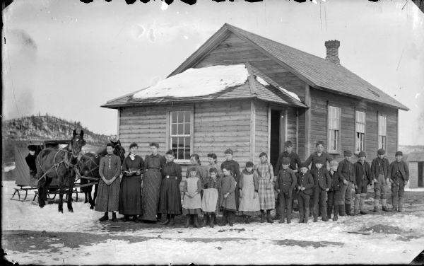 A group of four women and a group of children are standing in front of a building, perhaps a school. A man is sitting in a sleigh with two horses on the left.