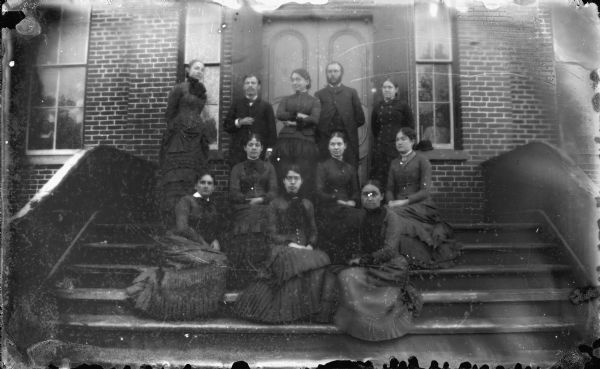 Two men and eight women wearing dark clothes are gathered for a group portrait on the entry stairs of a brick building. Identified as: Back row left to right: Connie Smith Hobart, unknown, Mary Wright, Frank Winter, unknown; Second row: unknown, Lizzie Parnell, unknown; Front row: unknown.