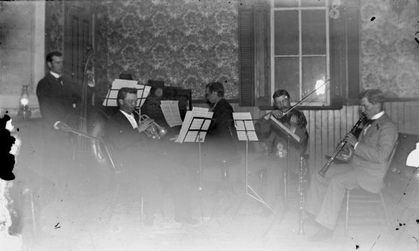 A group of musicians sitting indoors with their instruments and music stands. Includes a bassist, a trumpet player, a pianist, a violinist, and a clarinetist.