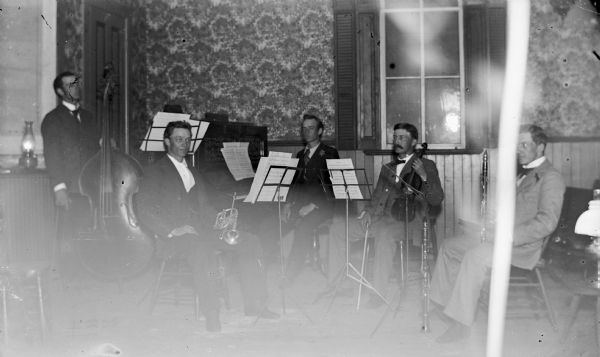 A group of musicians sitting with their instruments indoors. The five men are holding a bass, trumpet, piano, violin, and clarinet.