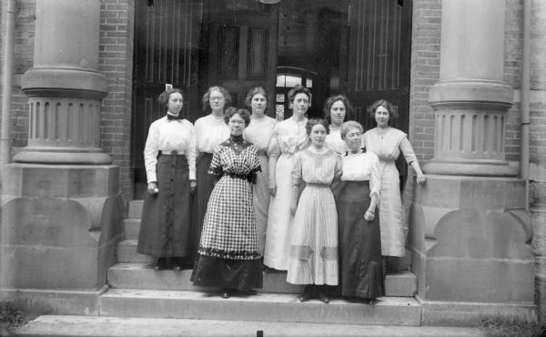 A group of nine unidentified women are standing on the front steps of a brick building.