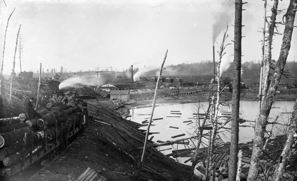 A view of logs transported by rail heading toward a group of buildings. A man is sitting on a stack of logs on the left. Other logs are floating in the water.