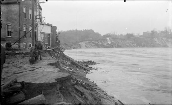 View along eroded shoreline of the damage from the 1911 flood of the Black River. A city street had been washed away. Three men are looking at the river.