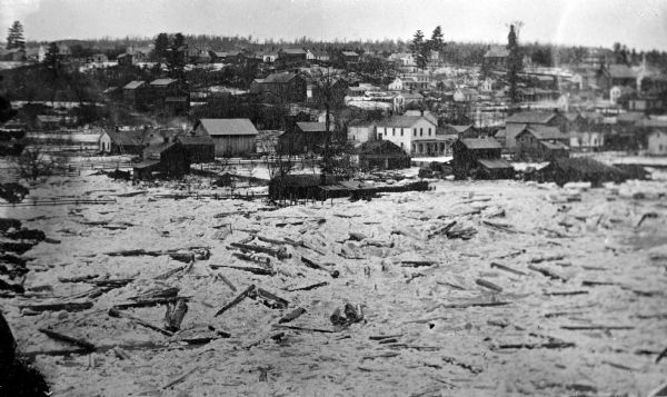 Elevated view of damage to buildings after the 1911 flood of the Black River. Wooden debris is floating in the flood water and encroaching the shoreline.