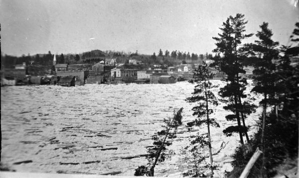 Elevated view across the high water on the Black River towards the town of Black River Falls. There is a large amount of wooden debris floating in the river.