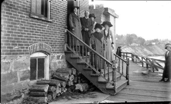 Outdoor group portrait of four women and a man standing on the back steps of a building. On the right is a woman standing on a wooden walkway with a railing that is along the river. In the background is a steep, eroded shoreline along the Black River.