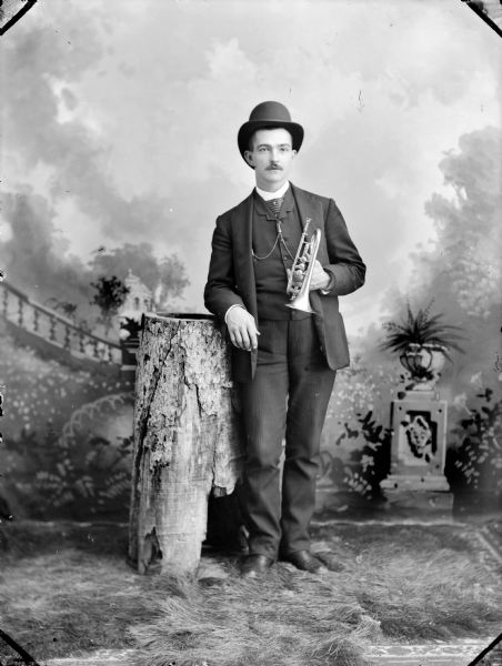 Full-length studio portrait in front of a painted backdrop of an unidentified man posing standing and holding a trumpet.