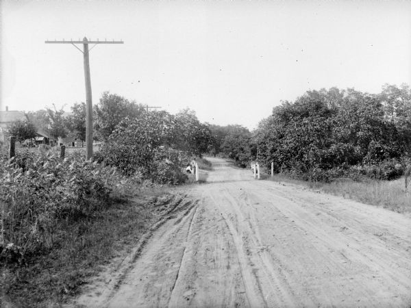 View down an unpaved country road lined with shrubs and short trees. A farm is on the left. Further down the road, four men are along both sides of the road near fence posts.