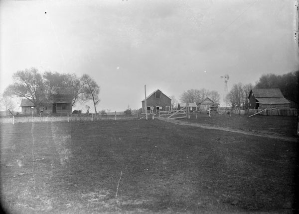 View across field towards a farm with a house, two barns, chickens, a windmill, and two smaller buildings.