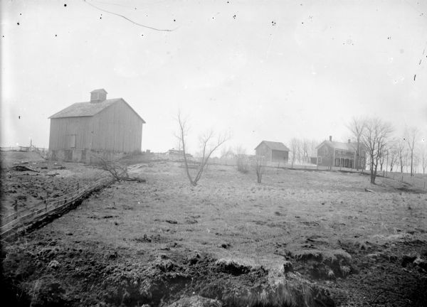 View across field towards a farm with a farmhouse, barn and stable.