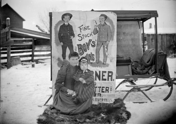 Outdoor portrait of a woman and girl sitting on a fur throw on the snow covered ground in front of a poster. The complete title on the poster is obscured, and reads, in part: "Fine Stock of Boys' __ing." A sleigh is parked behind the poster.