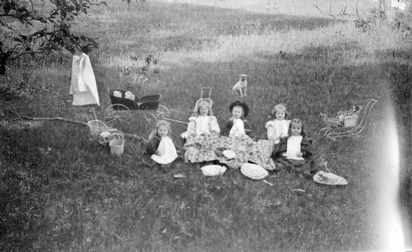 Group portrait of five girls sitting around a blanket in the grass. Each girl is wearing a napkin. Doll carriages and a dog are sitting behind them. A coat is hanging in a nearby tree on the left.