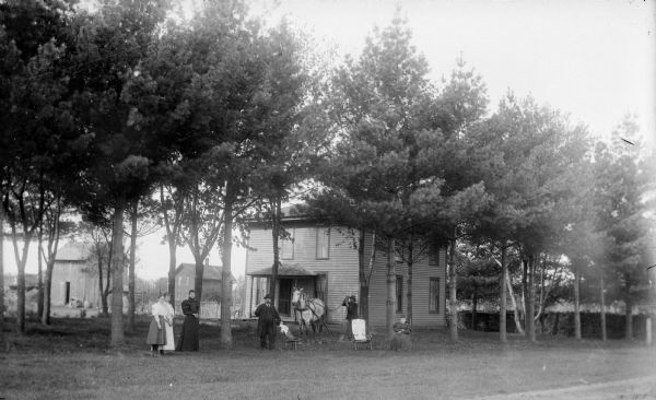 Four women, two men and a horse are posing in the front yard of a house. In the background are farm buildings.