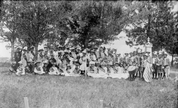Outdoor group portrait of girls wearing State sashes, and a group of boys on the right wearing uniforms. One boy has a drum, another boy has a flag, and the others have (wooden?) swords. Perhaps in a cemetery, with graves on the far right.