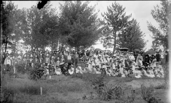 Outdoor group portrait of the participants of a patriotic pageant. The men are wearing suits, and some are wearing military medals. The boys are in uniform, with two boys holding drums. The girls are wearing state sashes, and the women, some with umbrellas, are wearing corsages.