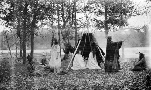 A group of eight people, men and women, are wearing Native American traditional dress. They are gathered near a tipi-like structure under trees. There is a river in the background, and leaves are covering the ground.