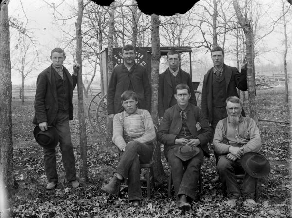 Outdoor group portrait of a group of seven men posing in front of C.R. Monroe's photographer's cart among trees.