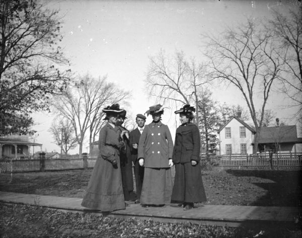 Outdoor view of four European American women and a man posing standing on the wooden walk in a town yard. Houses are in the background.