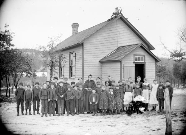 Outdoor group portrait of a one-room schoolhouse with the students and teachers posing in front for a group portrait. Snow is on the ground.