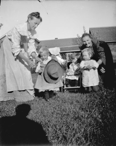 Outdoor view of two women and three small children standing in the yard of a house. One child is sitting in a stroller.