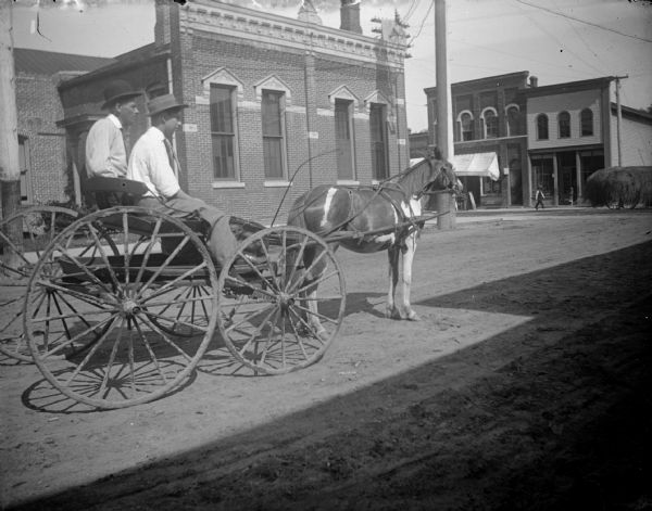 View towards two men posing sitting in a wagon pulled by a horse on a town road in front of a brick building. Location identified as south First Street.