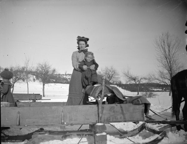 Outdoor view of a woman posing standing on a sled pulled by a horse. She is holding a small girl show is standing on the seat of the sled. Another girl is standing in the wagon on the far left.