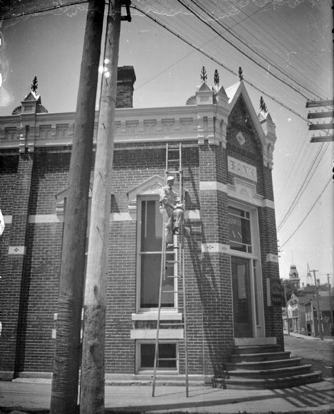Outdoor view of a man posing standing on a tall ladder propped up against a brick building identified as the Jackson County Bank at the intersection of First and Main Streets.