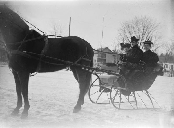 Outdoor portrait of a man and two women posing sitting in a sleigh pulled by a single horse on snow-covered ground. Buildings are in the background.