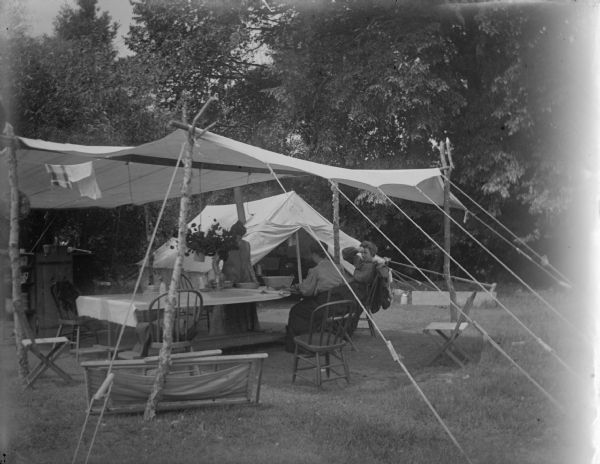 View towards a woman standing and two women sitting, at a table under a canopy and in front of a tent at a camp site.
