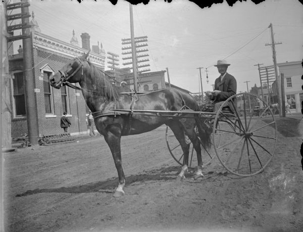 View of a man posing sitting in a buggy pulled by a single horse on an unpaved town street. Location identified as south First Street.