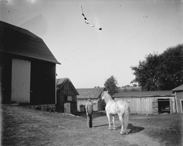 View of a man displaying a horse in the yard of several wooden buildings, possibly at a livery.