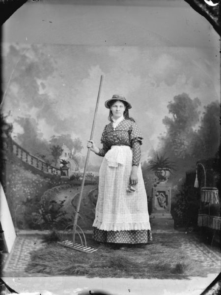 Full-length studio portrait in front of a painted backdrop of woman a posing standing and holding a rake with her right hand, and wearing a dark-colored dress and a light-colored apron.