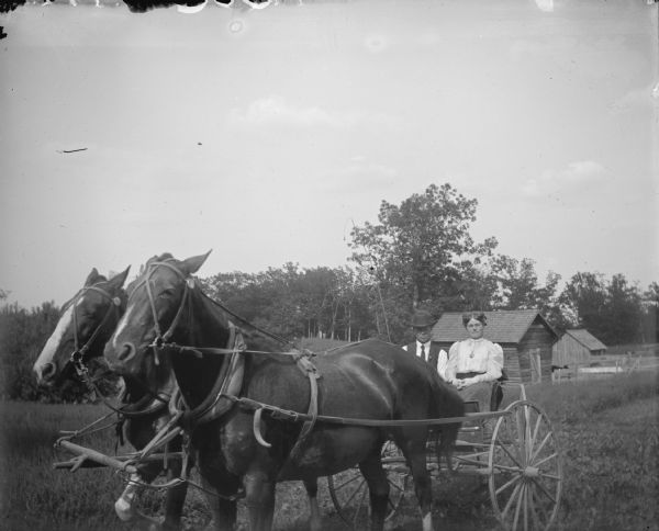 Outdoor view of a man and a woman posing sitting in a wagon pulled by a team of two horses. There are farm buildings in the background.