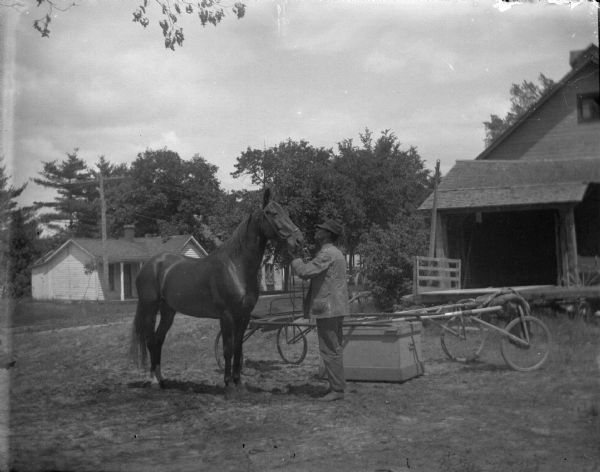 Outdoor view of a man displaying a horse near some racing harnesses in the yard of several wooden buildings.