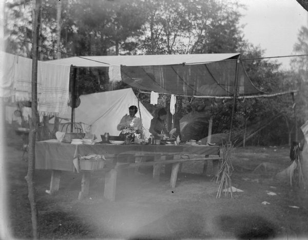 Main image: Outdoor view of two women standing at a table under a canopy in front of a tent at a camp site. Secondary image: Seen faintly in the awning area is a woman sitting in a chair.