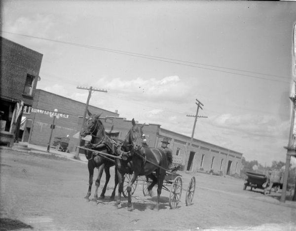 View towards a man sitting in a wagon pulled by a team of two horses on a town street. Location identified as a street in Black River Falls.