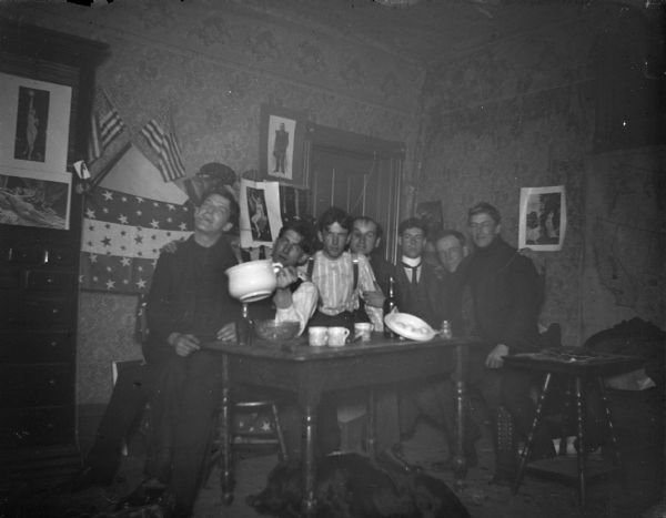 Group portrait of a group of men posing sitting and standing around a table. There are several female nude photographs hanging on the wall behind them.