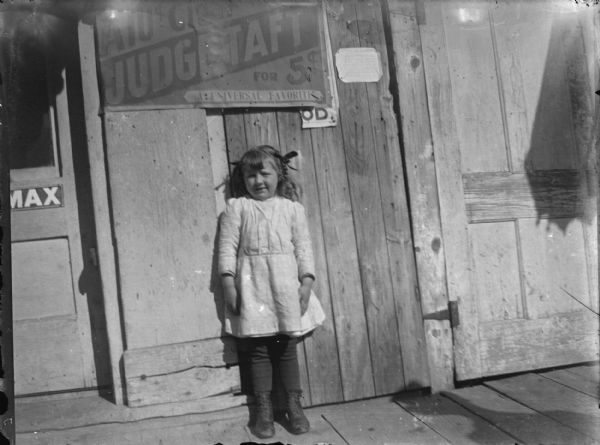 View towards a small girl posing standing on a wooden walkway against the front of a wooden building. Above her is a sign that reads, in part: "Judge Taft."
