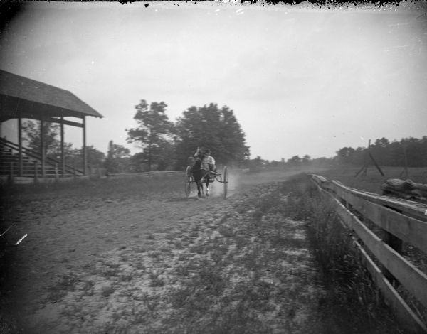 View down track towards a horse pulling a man in a racing buggy. Location identified as the racetrack at Jackson County Fairgrounds.