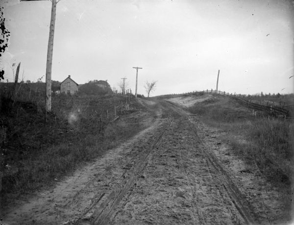 View down unpaved road.There is a fence along the road on the right, and a building in the distance on the left.