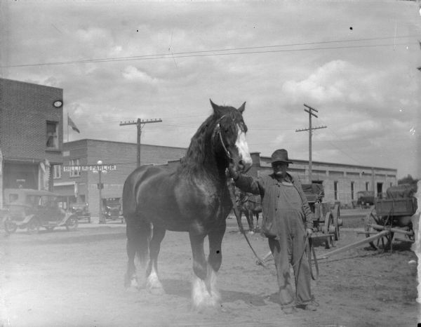 Outdoor view of a man posing standing and displaying a horse on a city street, with an automobile in the background on the left. A sign on the building behind the automobile reads: "Billiard Parlor."