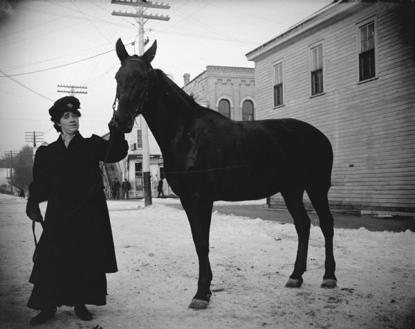 Outdoor view of a woman posing standing and displaying a dark-colored horse on a snow-covered town street. Pedestrians are on a street corner in the background. Location identified as south First Street.